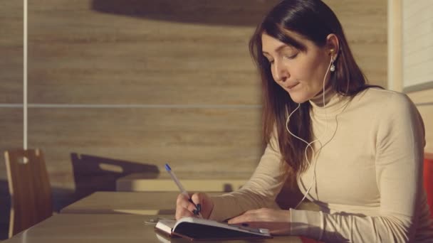 Jovem mulher sentada no café com fones de ouvido e fazendo anotações no caderno — Vídeo de Stock