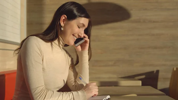 Mujer joven en un café hablando por teléfono y tomando notas en un cuaderno —  Fotos de Stock