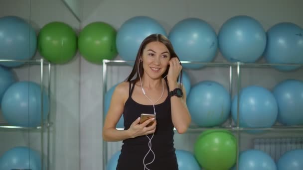 Mujer joven en el gimnasio con auriculares contra el fondo de bolas de gimnasia — Vídeos de Stock