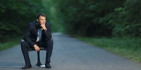 Homem bonito em um terno de negócios e fones de ouvido senta-se em uma cadeira em uma estrada florestal . — Fotografia de Stock