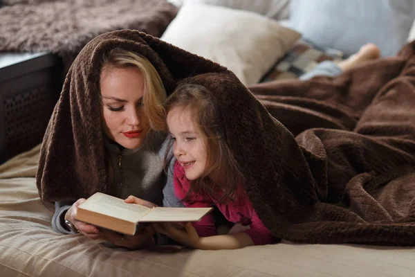Мать и дочь лежат на кровати под одеялом и читают книгу . — стоковое фото