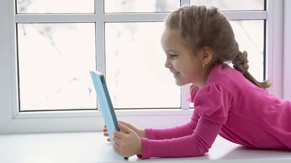 Geavanceerde meisje in een roze jurk zit bij het raam met een tablet — Stockfoto