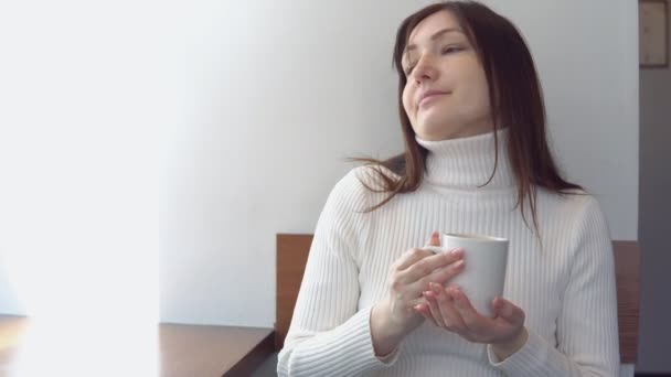 Ung kvinna i en vit tröja dricker kaffe på ett kafé — Stockvideo