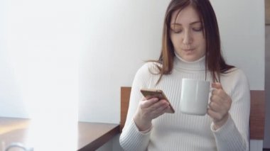 Kahve içme bir telefon ile beyaz kazaklı genç kadın