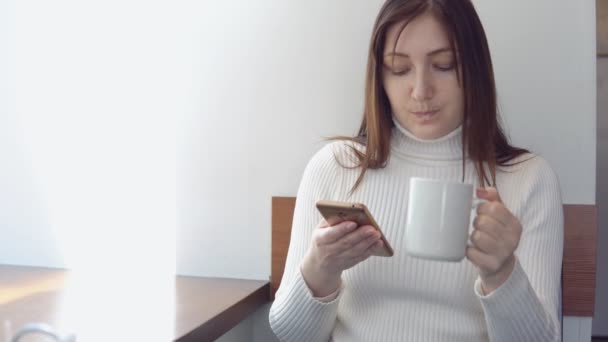 Giovane donna in un maglione bianco con un telefono che beve caffè — Video Stock