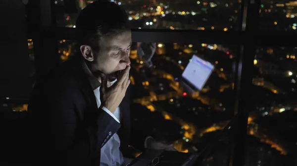 Tired sleepy man is working on his computer at night near the window with cityscape.