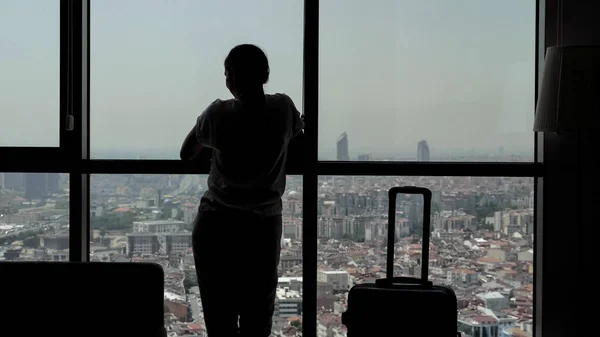 Silhouet van jong meisje reiziger met koffer komt binnen in hotelkamer met panoramisch uitzicht op de stad. — Stockfoto