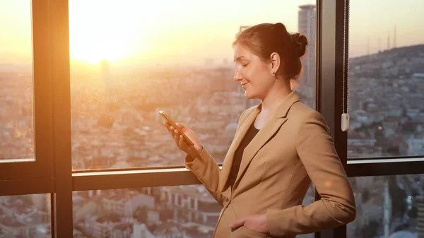 Jeune femme d'affaires au bureau parcourt le téléphone près de la fenêtre avec vue panoramique sur la ville . — Photo