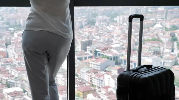 Vrouw met koffer in hotelkamer met panoramisch uitzicht op de stad, achteraanzicht. — Stockfoto