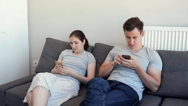 Pareja joven, hombre y mujer están mirando en sus teléfonos móviles sentados en el sofá. Dependencia de los aparatos . —  Fotos de Stock