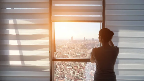 Kvinnan öppnar persienner, tittar på fönster med panoramautsikt över staden — Stockfoto