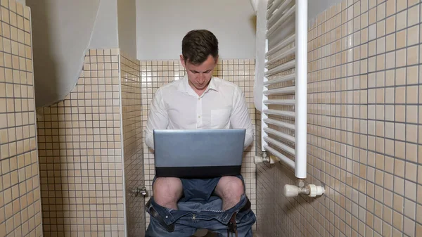 Молодой человек трудоголик работает в туалете на компьютере, печатая что-то и думая: . — стоковое фото