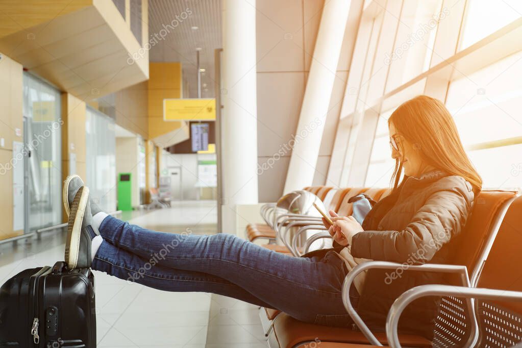 Young brunette woman in airport is waiting for her flight, something looking at smartphone.