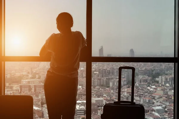 Силуэт юной девушки-путешественницы с чемоданом входит в гостиничный номер с панорамным видом на город . — стоковое фото