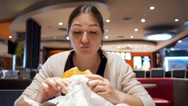 Young brunette woman is eating burger in fast food restaurant in food court. — Stock Photo, Image