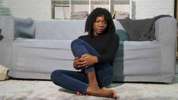Черная девушка в пуловере и джинсах сидит на белом полу у дивана — стоковое видео