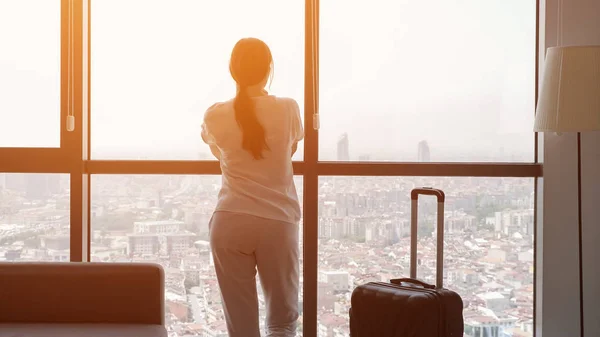 Jonge vrouw reiziger met koffer in hotelkamer met panoramisch uitzicht op de stad. — Stockfoto