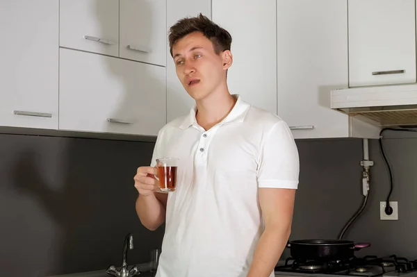 El joven soñoliento está tomando un té por la mañana en la cocina de su casa. . — Foto de Stock