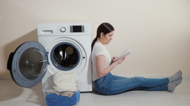 Upset woman with dirty laundry in box and manual in hands sits on floor — Stock Video