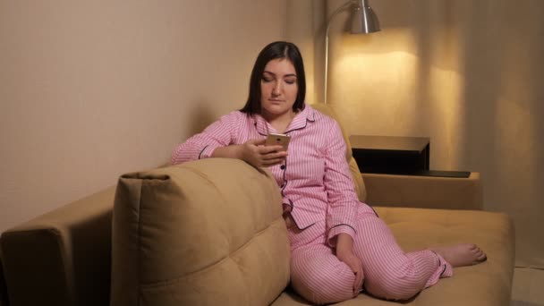 Eine Frau im Schlafanzug sitzt auf der Couch, ihr Handy im Hintergrund einer Lampe — Stockvideo