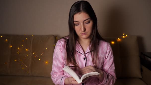 Una mujer morena por la noche se sienta en el sofá, lee un libro. Guirnaldas — Vídeos de Stock