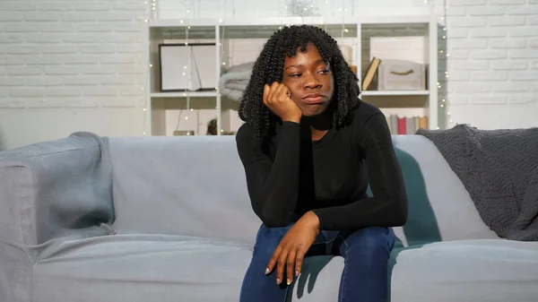 Aburrido negro chica se sienta en gris sofá contra blanco pared —  Fotos de Stock