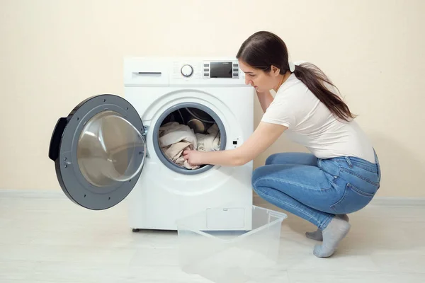 Женщина загружает грязную одежду в стиральную машину возле бежевой стены — стоковое фото