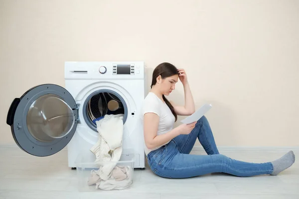 upset woman with dirty laundry in box and manual in hands sits on floor