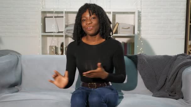 Красивая черная леди с вьющимися волосами сидит на сером диване и разговаривает — стоковое видео