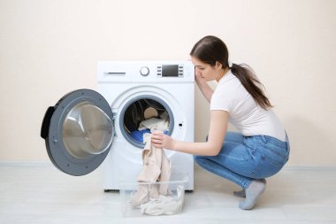 Kadın kirli çamaşırları bej duvarın yanındaki çamaşır makinesine dolduruyor.