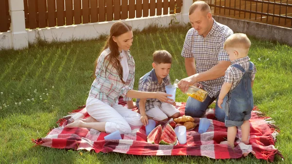 Šťastná rodina sedí na přikrývce a otec nalije pomerančový džus — Stock fotografie