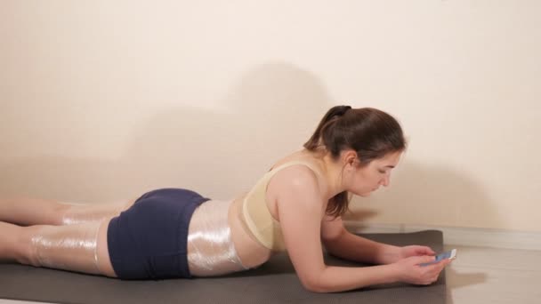 Mujer está acostada en una alfombra de gimnasio con un teléfono — Vídeos de Stock