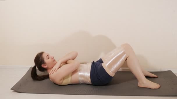 Eine junge Frau führt eine Übung auf der Matte aus und wickelt die Problemzonen mit Frischhaltefolie ein — Stockvideo