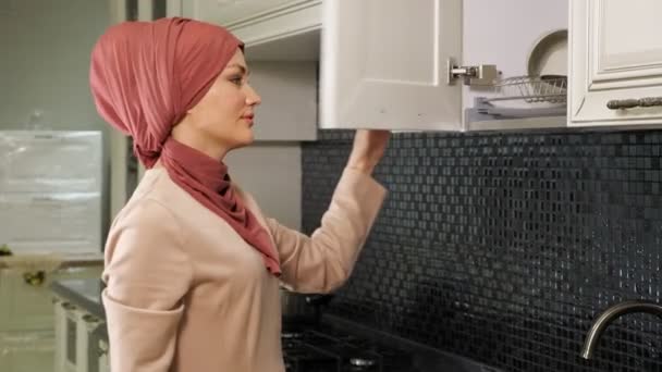 Женщина в хиджабе открывает кухонный шкаф и берет чистые тарелки — стоковое видео