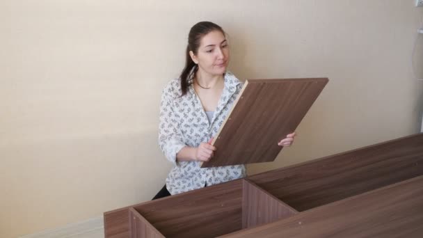 Девушка сидит на полу и пытается собрать части шкафа — стоковое видео
