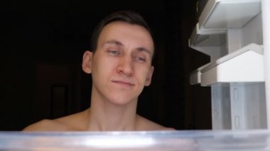 Uykulu adam buzdolabını açıyor esniyor ve yiyecek arıyor.