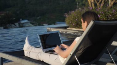 girl texts on laptop resting at hotel pool against bush