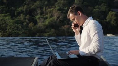 man talks on phone typing on laptop against forest and pool