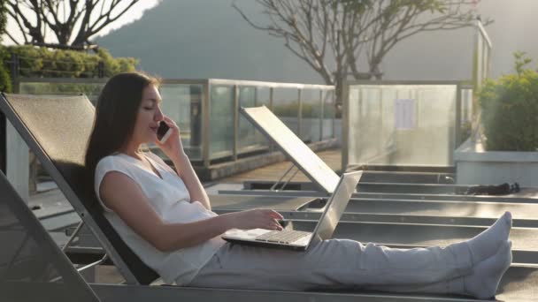 Chica habla por teléfono escribiendo en el ordenador portátil en el salón de descanso del hotel — Vídeos de Stock