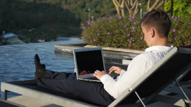 Бизнесмен работает над ноутбуком против леса и гостиничного бассейна — стоковое видео