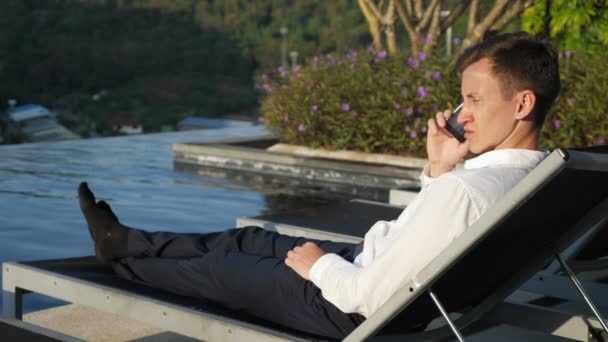 Guy talks on phone resting at hotel pool by bush at sunset — Stock video