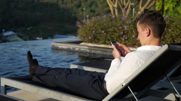 Man with phone rests on folding chair at pool against bush — Stock video