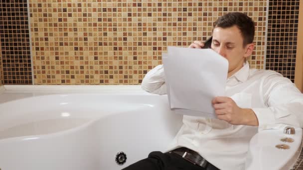 Мужчина в брюках и рубашке лежит в ванной и разговаривает по телефону — стоковое видео