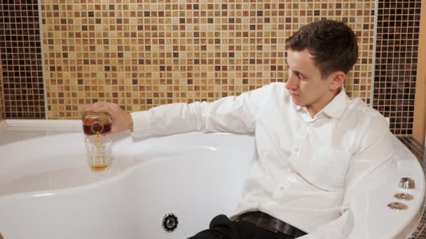 Мужчина в брюках и рубашке лежит в ванной и пьет алкоголь — стоковое видео