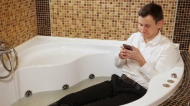 Pantolon giyen ve telefonlu tişörtlü bir adam su banyosunda yatıyor.