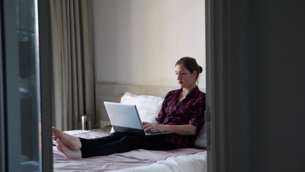Senhora senta-se na cama branca e tipos no laptop moderno cinza — Vídeo de Stock