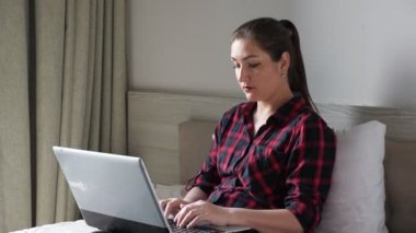 Güzel kız kahverengi yatakta oturur ve modern gri dizüstü bilgisayarda yazar.