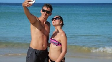 Gülümseyen mutlu çift gök mavisi okyanusun yanındaki güneşli sahilde selfie çekiyor.