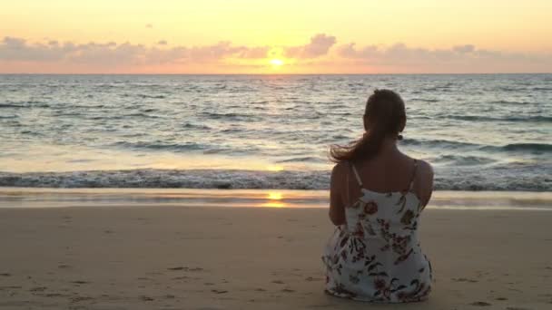 Mujer joven en vestido elegante se sienta en la playa al atardecer — Vídeos de Stock