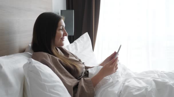 Dívka v županu studie on-line s tabletem v posteli hotelu — Stock video